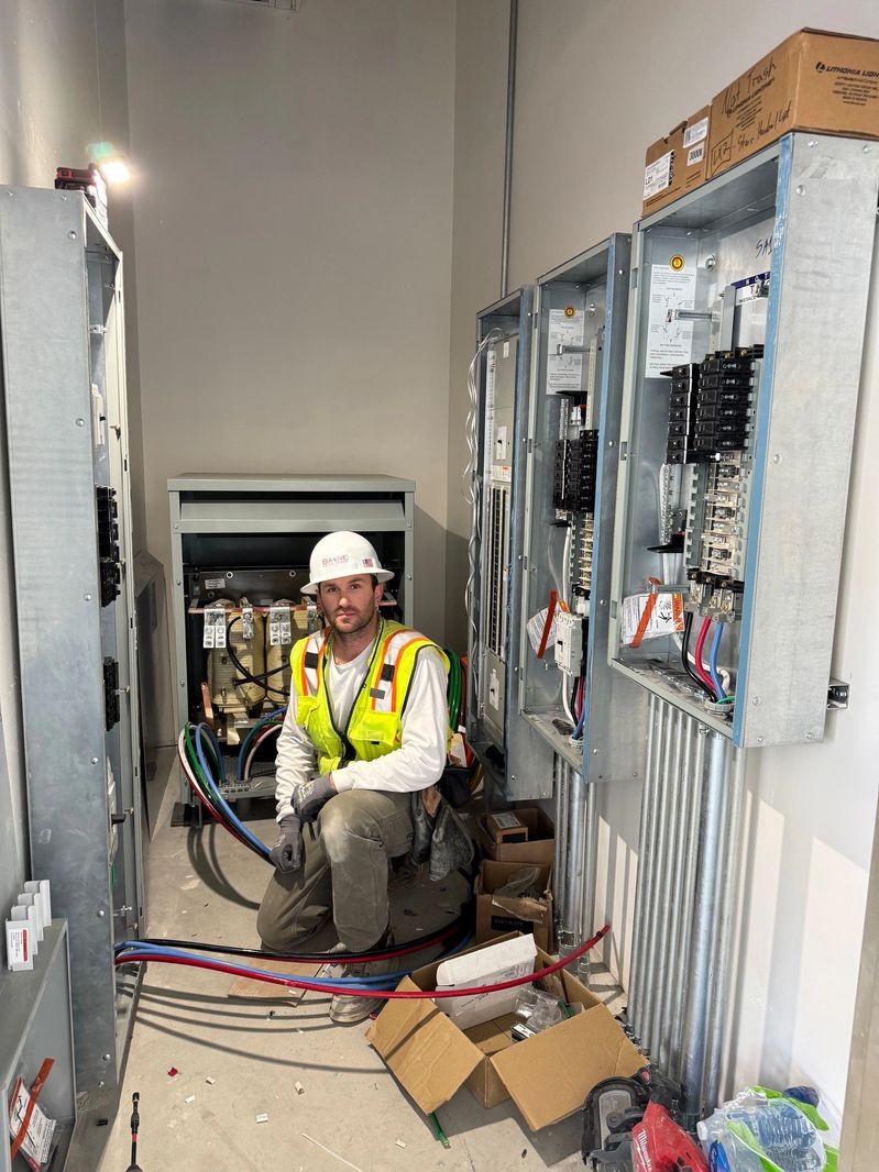 A Bayne Electrical electrician in a BAYNE hard hat working at open electrical panels with clean conduit runs